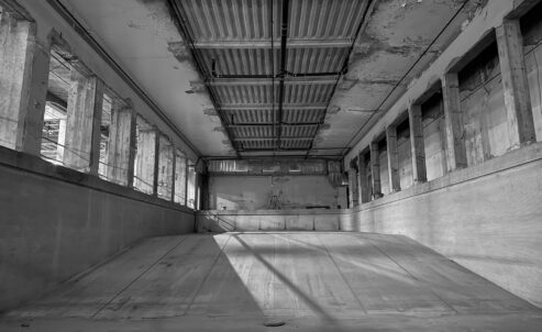 Black and white photograph of a cavernous concrete room with slanted floor, thick blocky columns, and exposed ductwork and pipes.