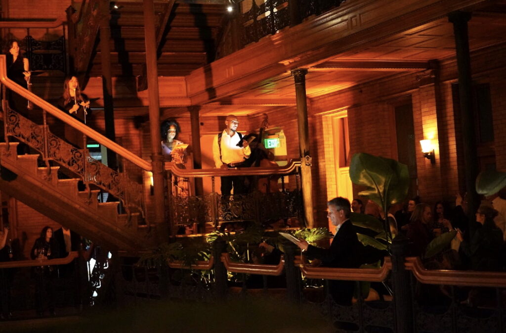 members of The Industry's company singing on the wrought iron stairs of the bradbury building with a warm glow and audience below in the shadows