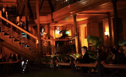 members of The Industry's company singing on the wrought iron stairs of the bradbury building with a warm glow and audience below in the shadows
